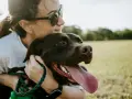 Ciemnobrązowy pies z wystawionym językiem, który szybko oddycha, przytulony do kobiety w okularach przeciwsłonecznych na tle zielonej łąki.